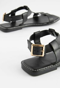 Mustat nahkasaappaat, joissa on litteä pohja, säädettävät hihnat ja kultasävyinen solki. Liukas pinta, jossa on valkoiset ompeleet.