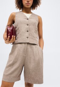 Γυναίκα με μπεζ αμάνικο γιλέκο με κουμπιά και matching σορτς μήκους μέχρι το γόνατο, κρατώντας μικρή μπορντό τσάντα στο ένα χέρι.