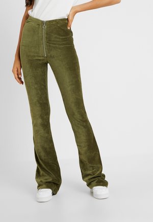 Frau trägt hochtaillierte Olivgrüne Cordhose mit Reißverschluss vorne, kombiniert mit weißen Turnschuhen, steht vor einem schlichten Hintergrund.