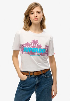Biała bawełniana koszulka z różowymi grafikami palm oraz niebieskim napisem "SUPERDRY". Krótkie rękawy i standardowy krój. Noszona z niebieskimi dżinsami.