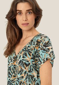 Kortärmad V-ringad topp i ett lätt material. Har ett flerfärgat tropiskt bladmönster i teal, svart och beige. Mjuk textur.