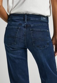 Jeans de mezclilla azul oscuro con cintura ajustada, piernas rectas, dos bolsillos traseros que presentan un patrón de costura ondulada y una etiqueta de la marca en la cinturilla.