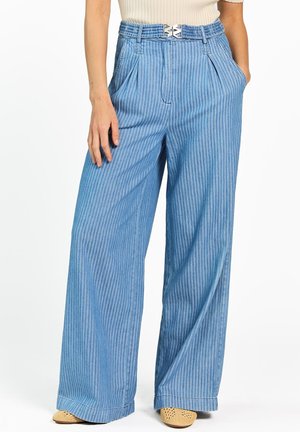 Vrouw draagt een blauw gestreepte broek met hoge taille en wijde pijpen, voorzien van plooien, voorzakken en een zilveren gesp, gecombineerd met beige gevlochten schoenen.