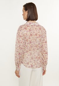 Transparante blouse met lange mouwen in lichtroze met een bloemenpatroon dat paarse en gouden accenten bevat, knoopmanchetten en een kraagnek.