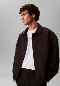 Smeđa, prevelika jakna s patentnim zatvaračem sprijeda, ukrasnim gumbima i širokim ovratnikom, slojevito nošena preko bijele majice s crnim logotipom.