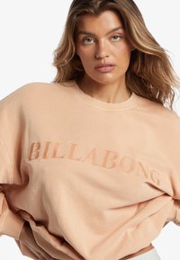 Mäkká mikina v broskyňovej farbe vyrobená z jemného materiálu, s tónovým vyšitou logom "BILLABONG" na prednej strane. Vo voľnom strihu s rebrovanými manžetami.