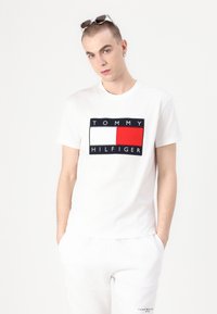 Balta medvilninė marškinėlių su apvaliu raudonu, baltu ir tamsiai mėlynu grafiniu elementu, kurio viduryje yra tekstas "TOMMY HILFIGER".