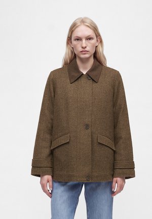 Ung kvinde iført en brun fiskebenfrakke med stor krave og klaplommer, kombineret med lyseblå jeans, stående mod hvid baggrund.