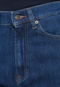 Jeans de ganga azul escuro com costuras laranja. Apresentam fecho de botão e fecho éclair, passadores para cinto e um bolso frontal com rebite.