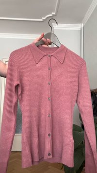 Rožinė raukinėta ilgų rankovių kardiganas su apykakle ir sagomis priekyje, pakabinta ant pilkos pakabos patalpoje.