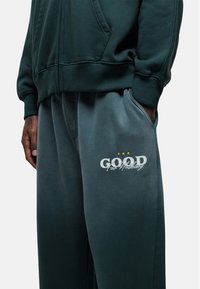 Donkergroen zip-up hoodie gecombineerd met grijze sweatpants met een witte bedrukte logo en ontwerp, gemaakt van een zachte katoenmix.