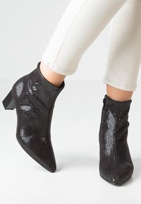 Bottines noires avec un motif en cuir de serpent texturé, bout pointu et un talon épais. Équipées d'une fermeture éclair latérale pour un enfilage facile.