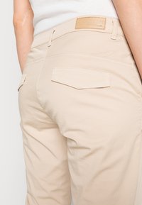 Calças de algodão bege com cintura média, bolsos traseiros com abas anguladas e um acabamento suave com detalhes de costura mínimos.
