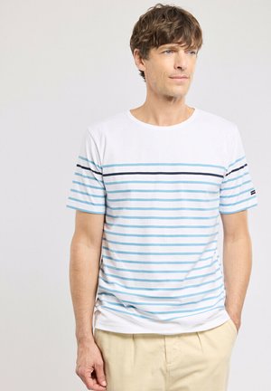 Homme portant un t-shirt blanc à manches courtes avec des rayures horizontales bleu clair et bleu marine, pantalon beige, debout devant un fond clair uni.