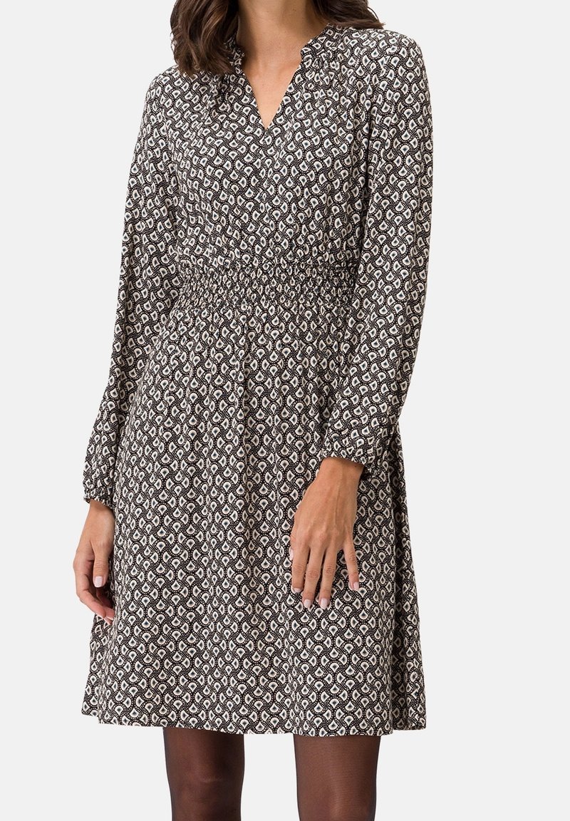 Femme portant une robe à manches longues à motifs noir et blanc avec taille froncée et col en V sur un fond uni.