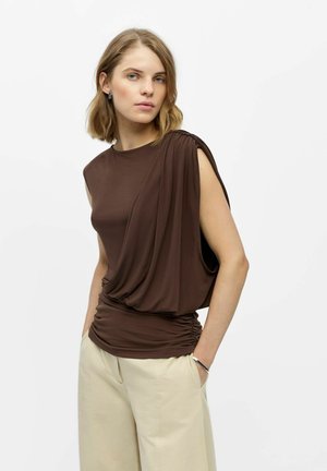 Femme aux cheveux blonds courts portant un haut marron sans manches drapé et un pantalon beige à jambes larges, debout les mains dans les poches devant un fond blanc.