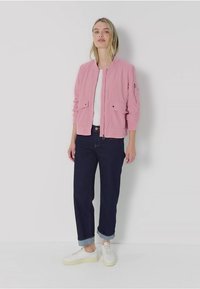 Jasnoróżowa kurtka bombowca z zapięciem na suwak, z dwiema przednimi kieszeniami, w połączeniu z ciemnoniebieskimi spodniami z podwiniętymi rękawami i białymi trampkami.