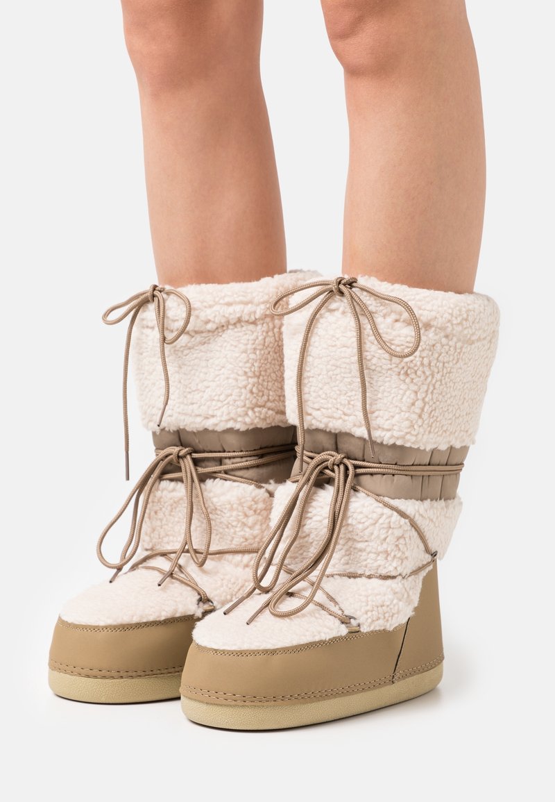 Jambes portant des bottes d'hiver beige et crème doublées en sherpa, avec des semelles épaisses et plusieurs lacets noués autour de la cheville et du mollet.