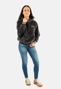 Grijze fleece hoodie met een voorzak en witte gestreepte boorden, gecombineerd met slanke blauwe jeans en beige sneakers met bordeauxrode accenten.