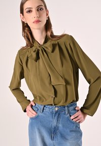 Olivgrüne Bluse mit Schleife am Kragen, langen Ärmeln und gepufften Details, kombiniert mit einem hellblauen, hochgeschnittenen Jeansrock.