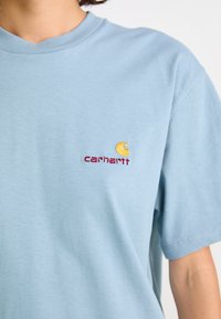 Svetlomodré tričko s malým vyšitým logom Carhartt v žltej a červenej farbe na hrudi, nosené osobou.