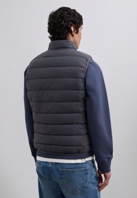 Hombre vestido con un chaleco acolchado gris oscuro sobre un sudadera marinera y jeans azules, visto desde detrás contra un fondo claro y liso.