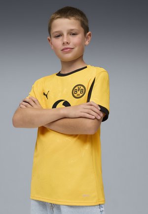 Geel voetbalshirt met zwarte accenten, heeft een subtiel patroon, ronde hals, en logo op de borst. Korte mouwen, sportief ontwerp.