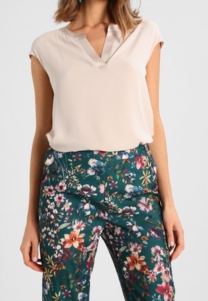 Frau trägt eine ärmellose beige Bluse, die in türkisfarbene Hosen mit buntem Blumenmuster gesteckt ist, steht mit entspannten Händen an den Seiten.