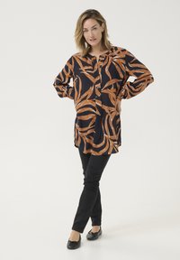 Tunique noire à manches longues avec un motif en vagues oranges, fermeture à boutons, fentes sur les côtés, assortie à un pantalon slim noir et des ballerines noires.