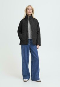 Sort kviltet jakke med høj krave, knappelukninger, båret over en grå sweater, parret med bukser i vidde i blå denim og grå sko.