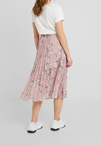 Kobieta ubrana w biały t-shirt w paski, różową spódnicę w kwiaty o plisowanym kroju do midi i białe, masywne sneakersy, stojąca na tle jednolitego szarego tła.