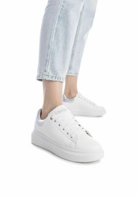 Zapatillas blancas hechas de material sintético, con una textura suave, punta redondeada y una suela gruesa. Diseño con cordones y acentos mínimos.