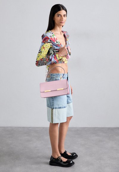 Borsa in pelle rosa con accenti dorati, forma rettangolare e manico a catena; indossata con una maglietta stampata colorata e pantaloni di jeans corti.