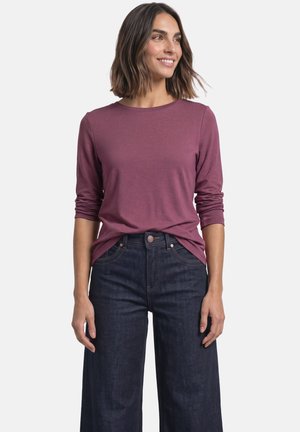 Kvinde smilende iført en langærmet støvet lilla top og mørke, højtaljede, vide jeans mod en ensfarvet hvid baggrund.