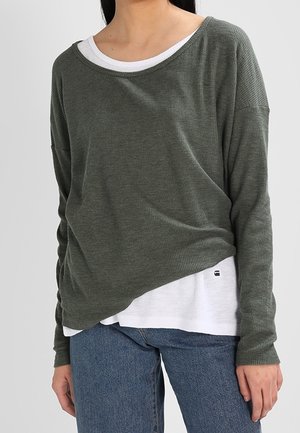 Femme portant des chemises longues superposées vertes et blanches avec un jean bleu sur un fond uni.