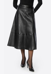 Jupe en cuir noir à design évasé, avec des coutures et une texture lisse. Associée à des chaussures à talons hauts noires et des collants transparents.