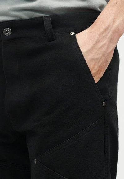 Χέρι στην μπροστινή τσέπη μαύρων παντελονιών με ορατή ραφή, καρφιά και μεταλλικό κουμπί στο ύψος της μέσης.