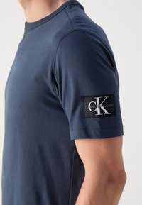 Mies, který носí темно-синюю футболку с коротким рукавом и черно-белым патчем логотипа Calvin Klein Jeans на верхней руке.