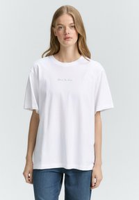 Maglietta bianca in cotone con maniche corte e scollatura rotonda, caratterizzata da un testo ricamato blu sottile sul petto. Design semplice e vestibilità ampia.