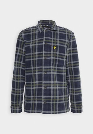Camicia di flanella a quadri blu navy con linee verdi e grigie, dotata di chiusura con bottoni, colletto e un piccolo emblema giallo sul lato sinistro del petto.