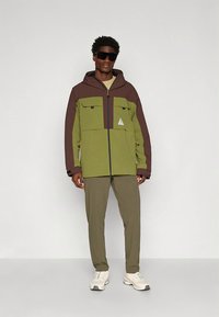 Vyras, vilkintis žalią ir rudą gobtuvu su gaubtu striukę, alyvuogių spalvos kelnes, baltus sportinius batelius ir dideles juodas saulės akinius, stovintis priešais šviesią, paprastą foną.