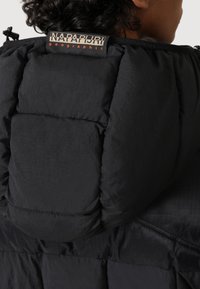 Osoba ubrana w czarną pikowaną kurtkę Napapijri Geographic z kapturem, z widoczną metką na karku.