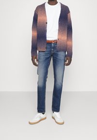 Pletený svetr v gradientních odstínech fialové a oranžové, s knoflíky, nošený přes bílé tričko a doplněný modrými džínami a bílými teniskami.