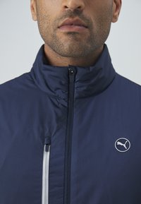 Giacca blu navy con colletto alto, chiusura a zip e logo Puma argentato. Realizzata in un materiale leggero e liscio con una finitura testurizzata.