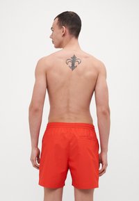 Pantaloni scurți de înot roșii, cu talie elastică, buzunare laterale și un buzunar pe spate. Modelul prezintă un tatuaj pe partea superioară a spatelui. Design minimalist.