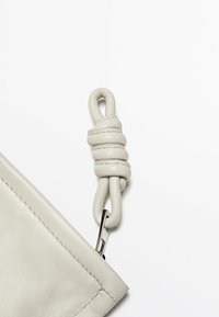 Pochette in pelle grigia chiara con tracolla annodata e hardware argentato. Presenta una superficie liscia e linee pulite, con dettagli di cucitura.