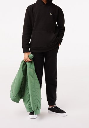 Person trägt schwarzen Hoodie und schwarze Hose, hält eine grüne Bomberjacke, steht mit einer Hand in der Tasche und trägt schwarze Sneakers mit weißen Sohlen.