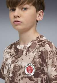 Brūns un bēšs rakstains T-krekls ar apaļu kakla izgriezumu, kurā ir FC St. Pauli logo un Puma simbols kreisajā pusē.