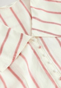 Chemise en lin blanc à rayures verticales roses, boutons recouverts de tissu et une étiquette indiquant qu'elle contient du lin.