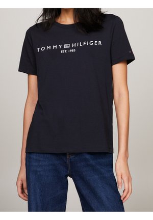 T-shirt noir Tommy Hilfiger avec texte de logo blanc "TOMMY HILFIGER EST. 1985" porté avec un jean bleu sur une personne devant un fond neutre.